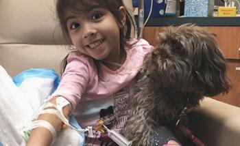 A smiling girl having blood drawn and holding a dog in her lap