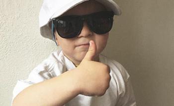 A young boy wearing an oversized cap and sunglasses giving a thumbs up.